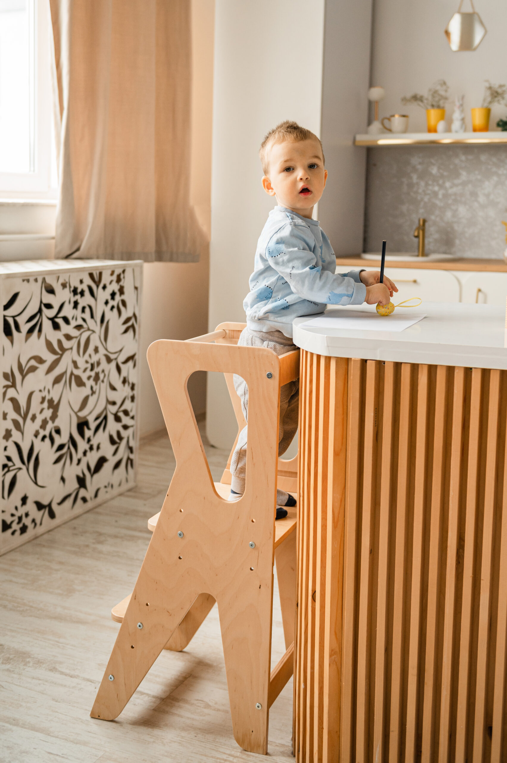 A toddler in blue pajamas sits on a wooden step stool at the kitchen counter, holding a spoon and a yellow bowl.