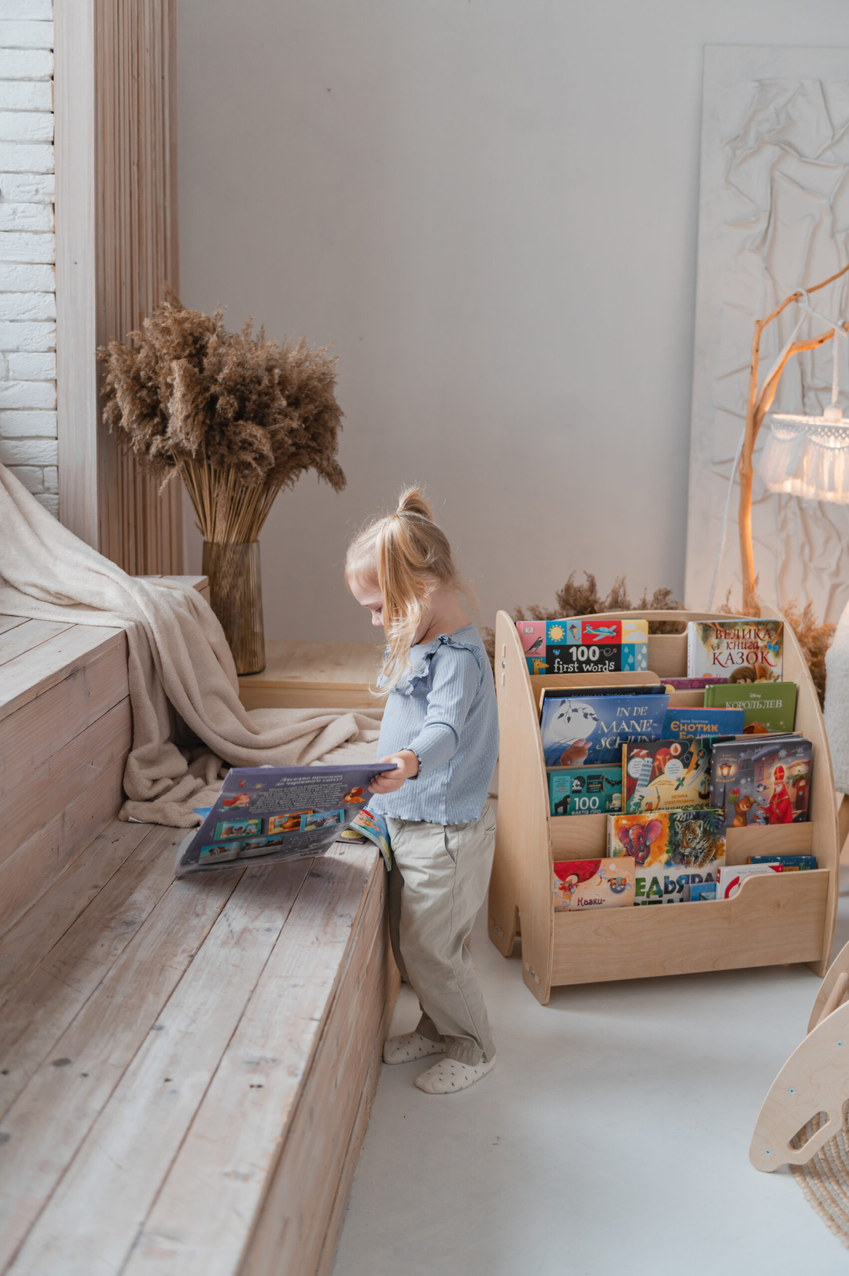 A young girl stands next to a wooden step and looks at a book on a bookshelf filled with children's books.