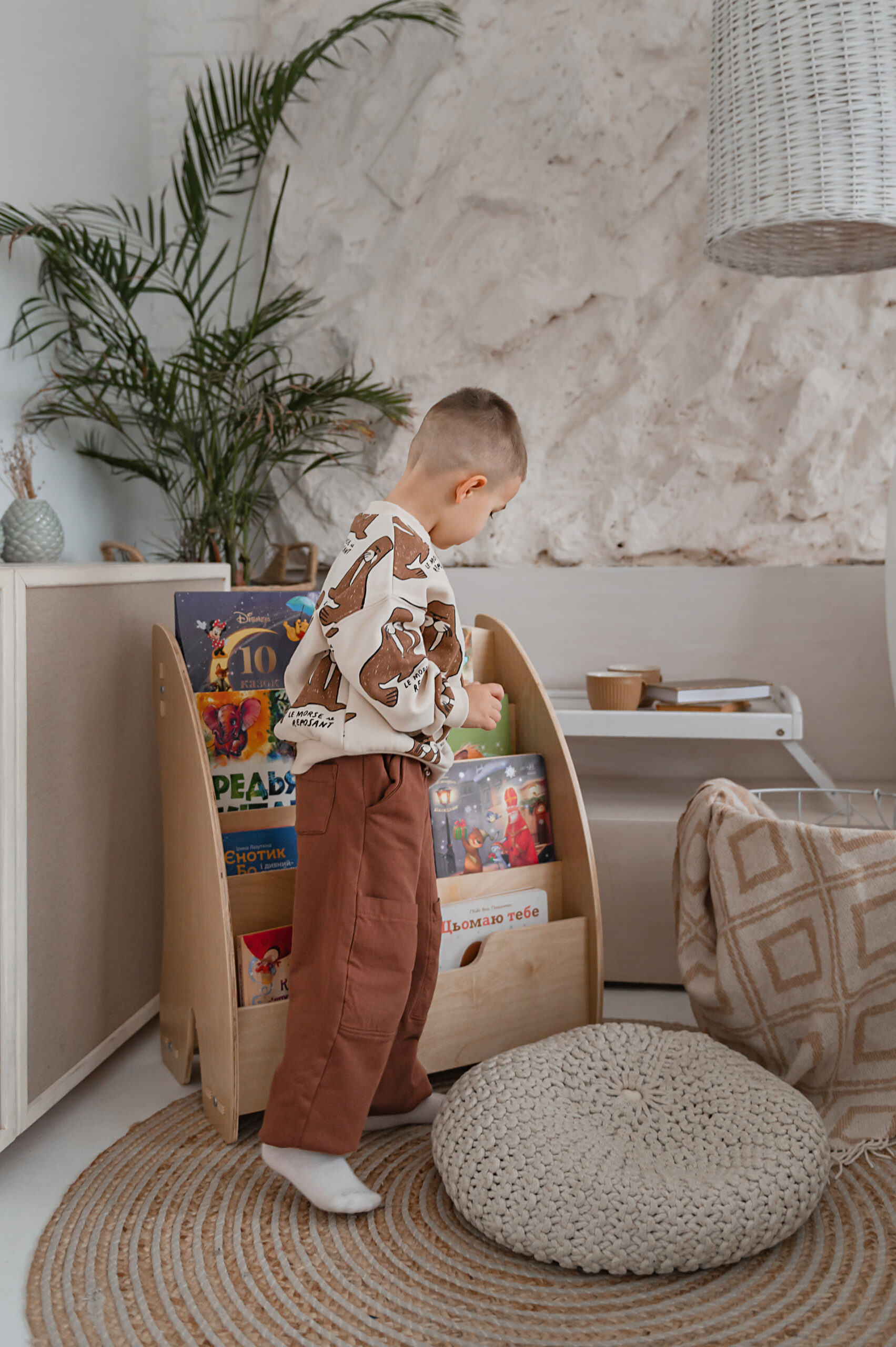 A small child in brown pants selects a book from a wooden bookshelf in a cozy, modern room with neutral tones.