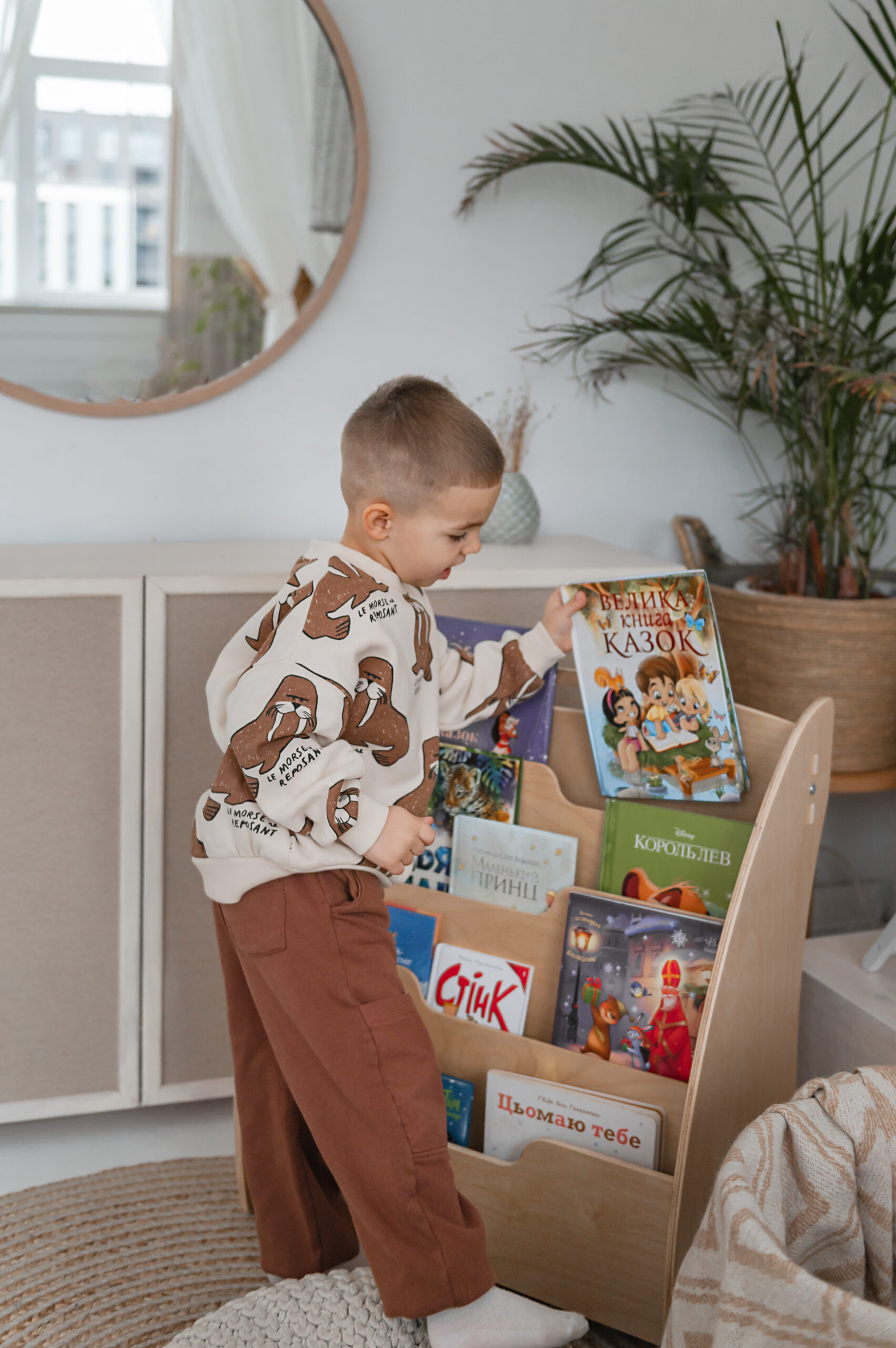A little boy takes a colorful picture book from a wooden bookshelf in a cozy, modern room with plants.