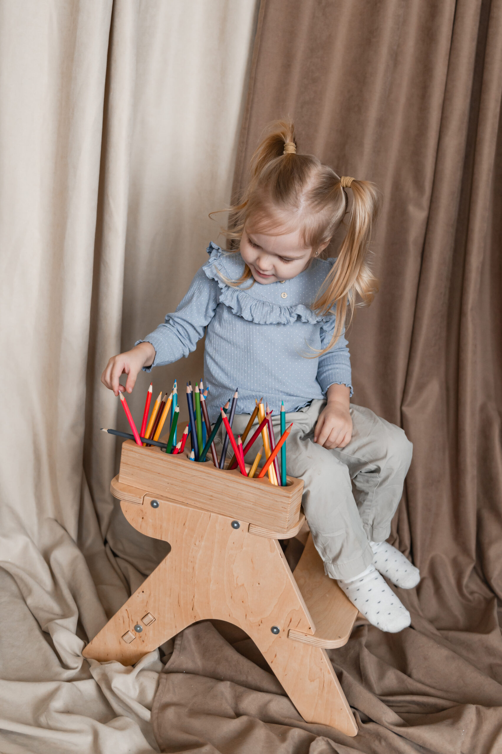 A young girl sits on a wooden chair, holding a box full of colorful crayons, smiling and looking down.