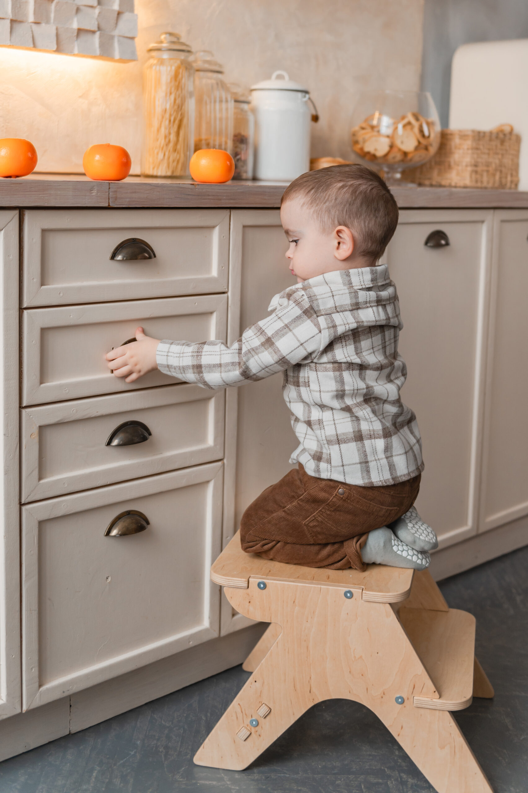 A toddler kneels on a wooden step stool, reaching out to open a kitchen drawer next to oranges on the countertop.
