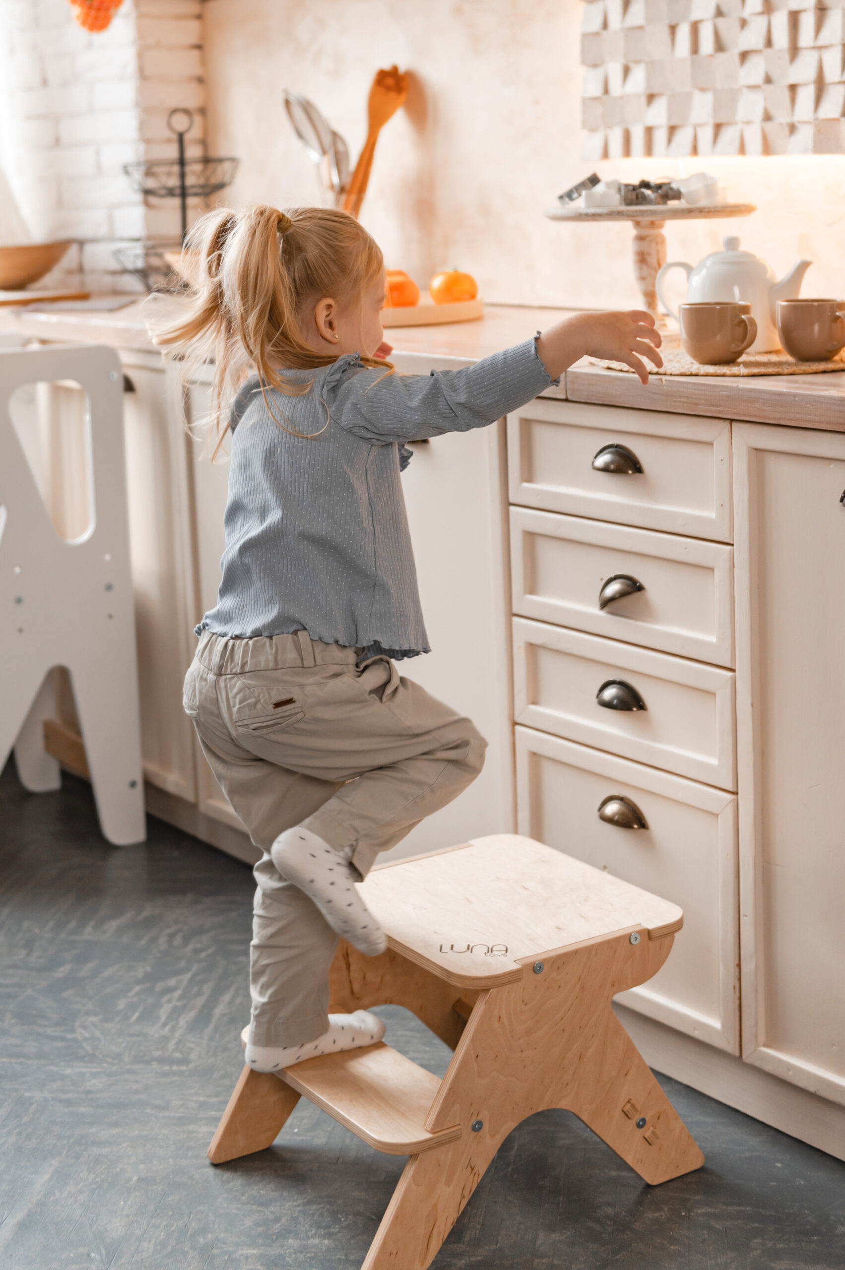A small child climbs onto a wooden stool in the kitchen, reaching out to the countertop where cups and a teapot are placed.
