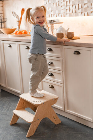 A smiling girl stands on a wooden step stool in a kitchen, reaching for a teapot on the countertop.