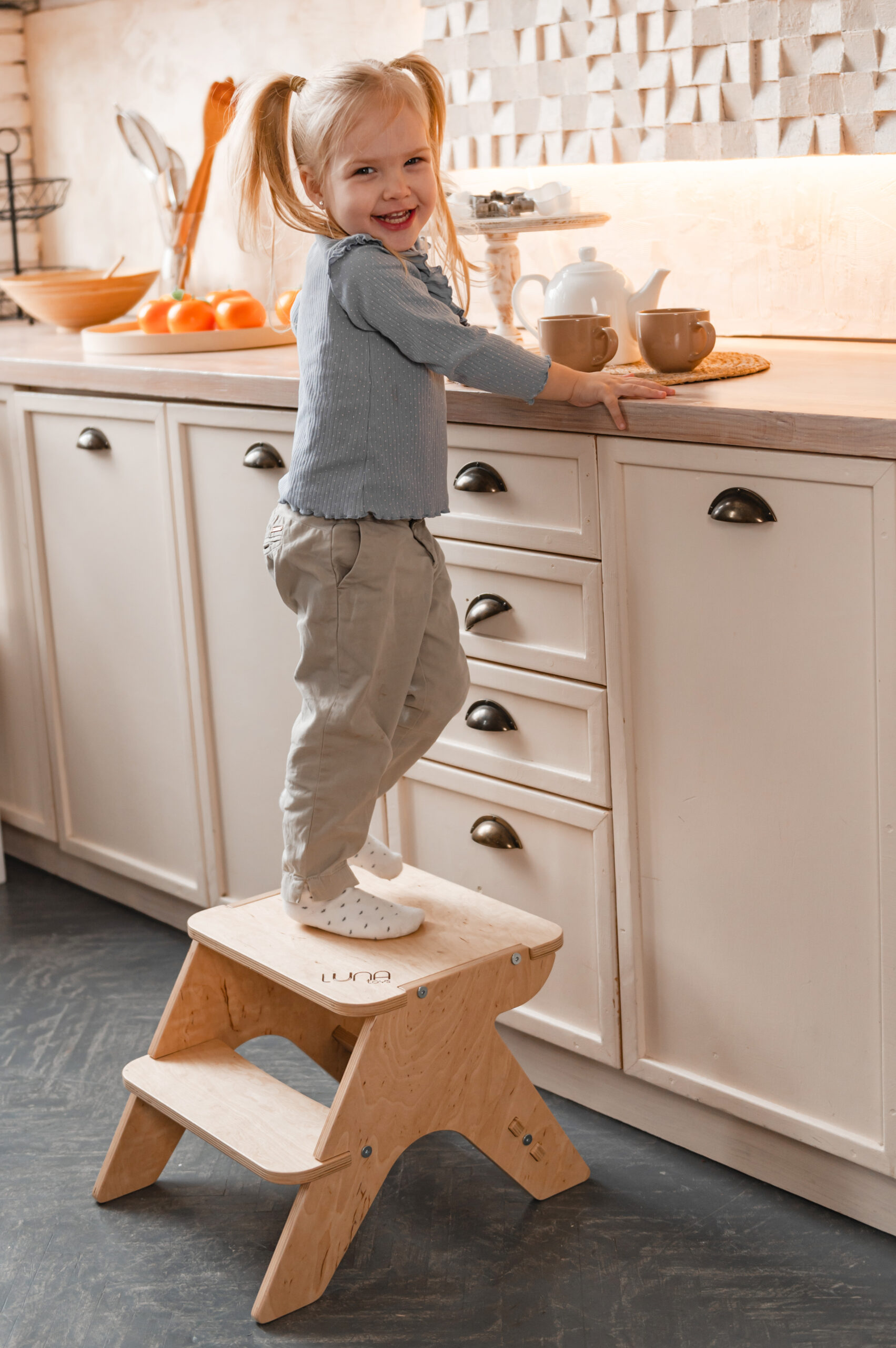 A smiling girl stands on a wooden step stool in a kitchen, reaching for a teapot on the countertop.
