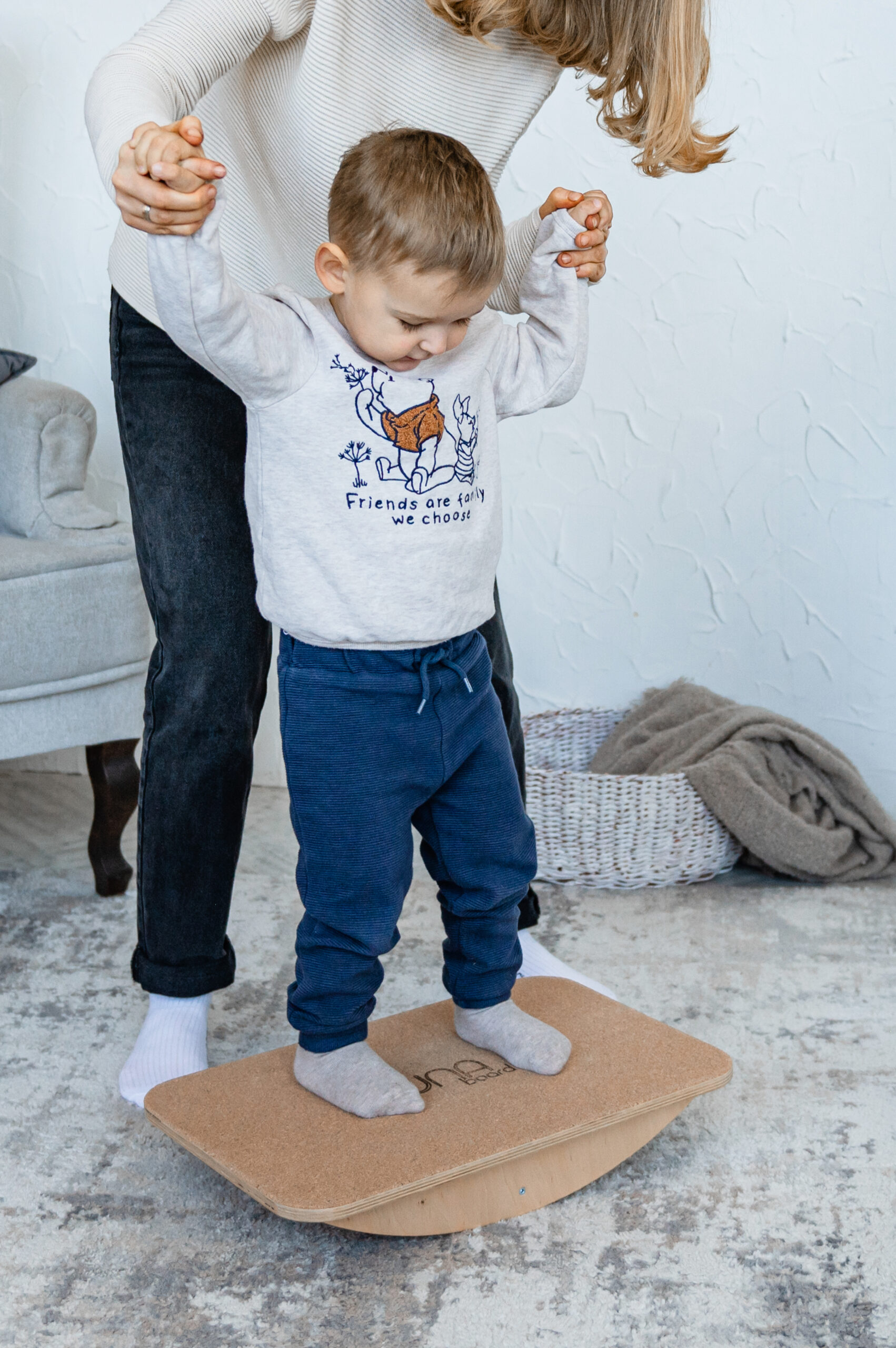 An adult helps a small child maintain balance on a wooden rocking board indoors by holding the child's hands.