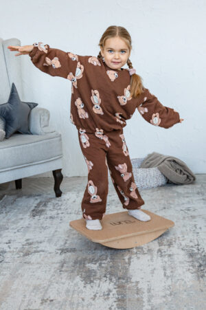 A little girl in brown pajamas balances on a wooden board in the living room, extending her arms to keep her balance.