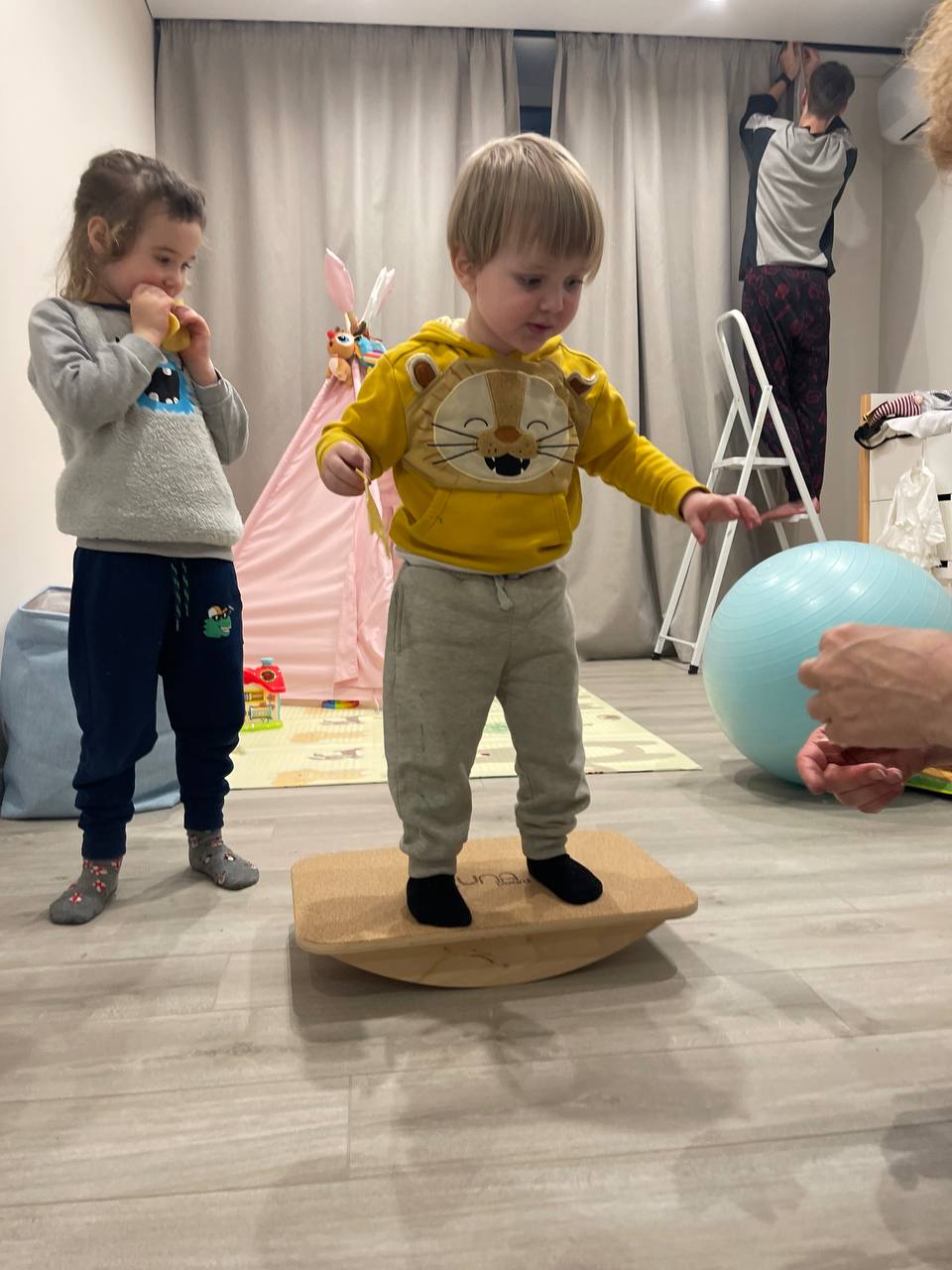 A toddler balances on a wooden board, a girl watches nearby, and an adult reaches out a hand - in a cozy room.
