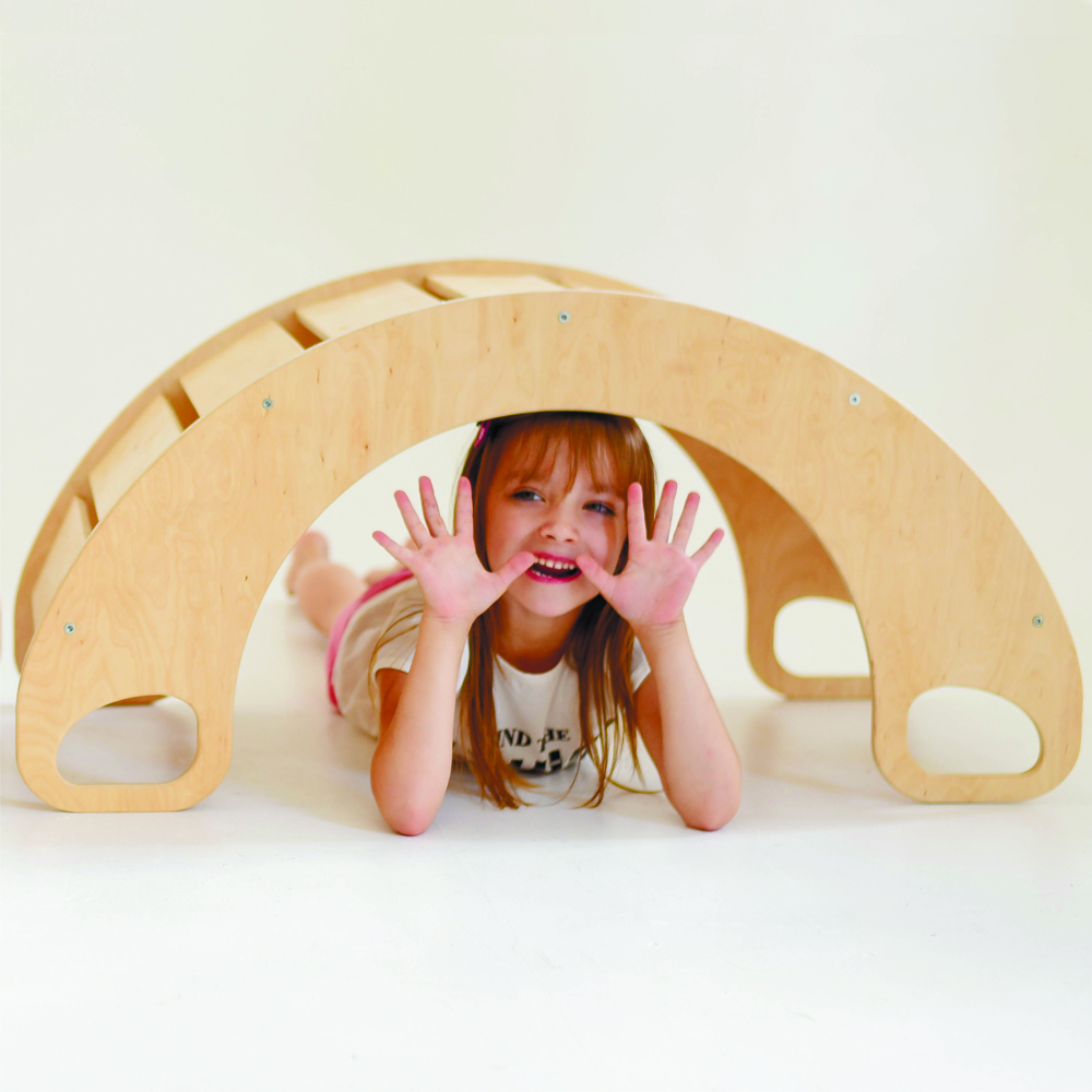 A smiling girl lies under a wooden arch, holding her hands up and looking through the opening.