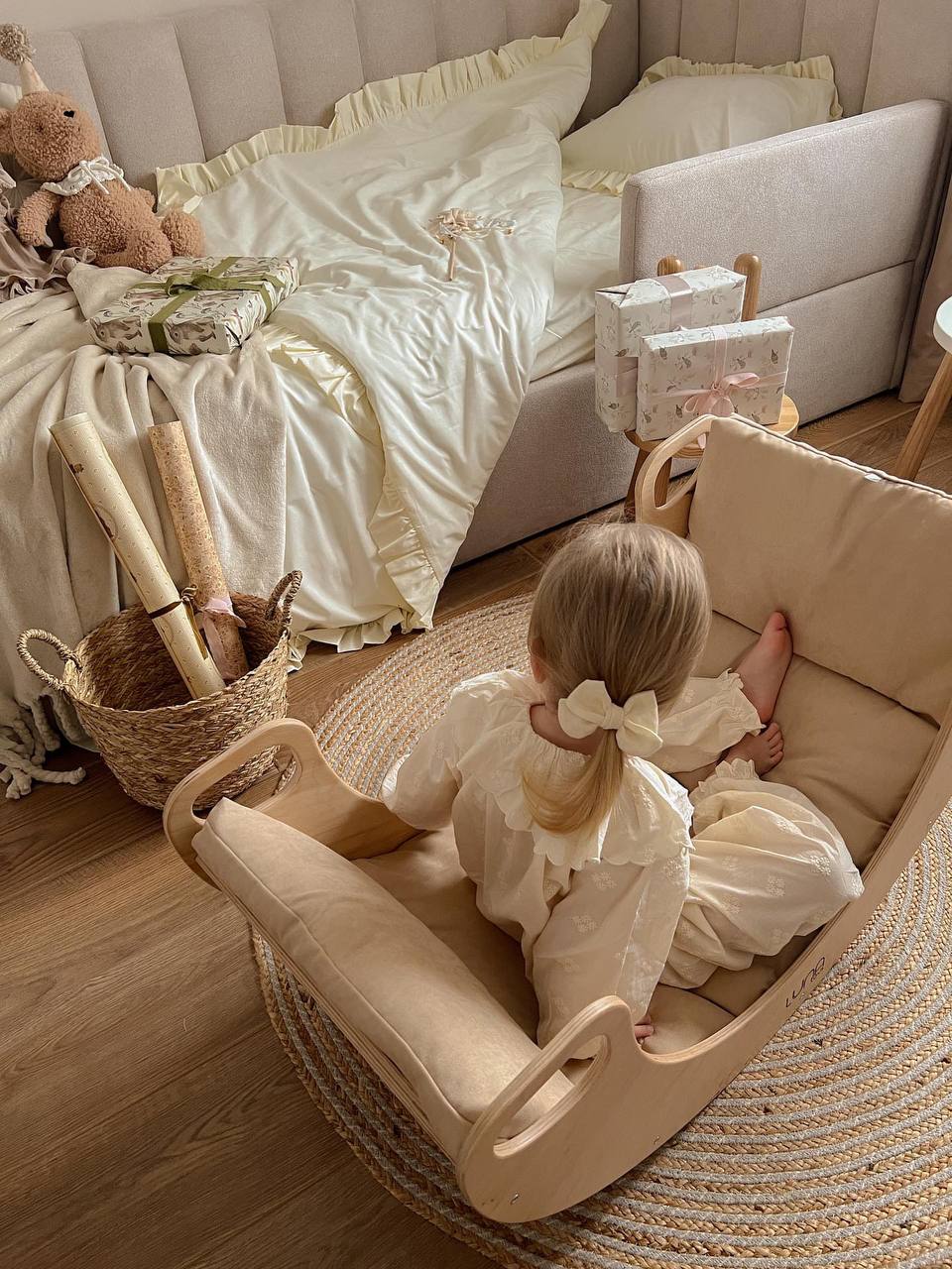 A small child in a cream-colored dress sits in a wooden rocking chair on a cozy rug in a softly lit, tidy bedroom.