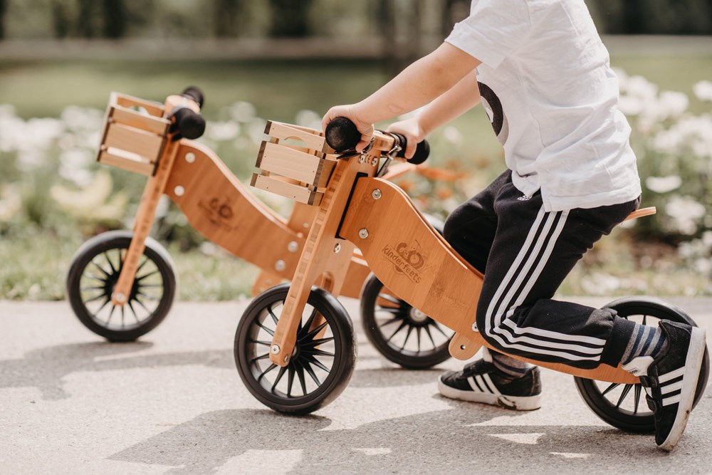 A child rides the Kinderfeets 2-in-1 Wooden Balance Bike Tiny Tots Plus Bamboo outdoors on a sunny day.