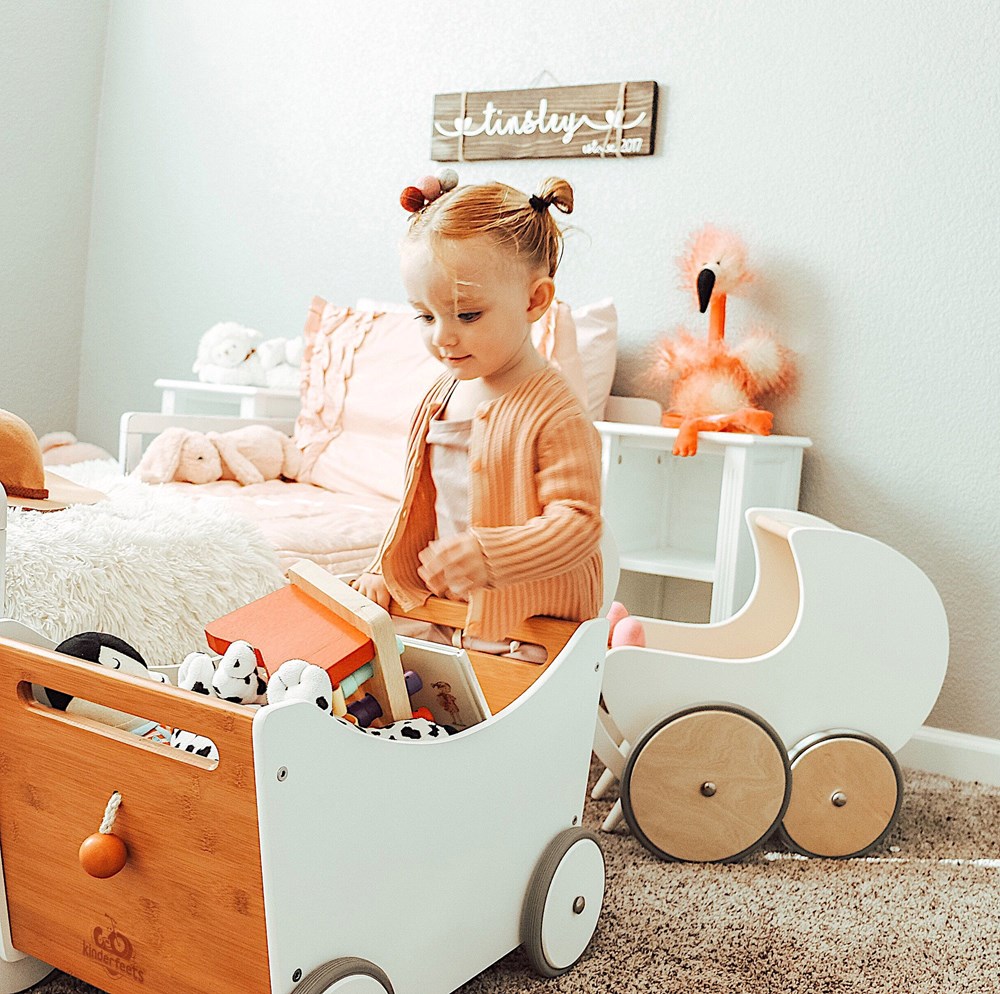 Ein Kleinkind spielt in einem gemütlichen, in Pastelltönen gehaltenen Zimmer mit der „Kinderfeets 2-in-1 Spielzeugkiste aus Holz, weiß“, die mit Spielzeug gefüllt ist.