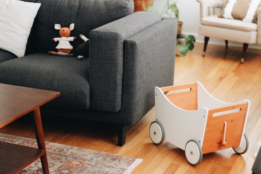 Die weiße „Kinderfeets 2-in-1-Spielzeugkiste aus Holz“ steht auf einem Holzboden neben einem grauen Sofa und einem Couchtisch.