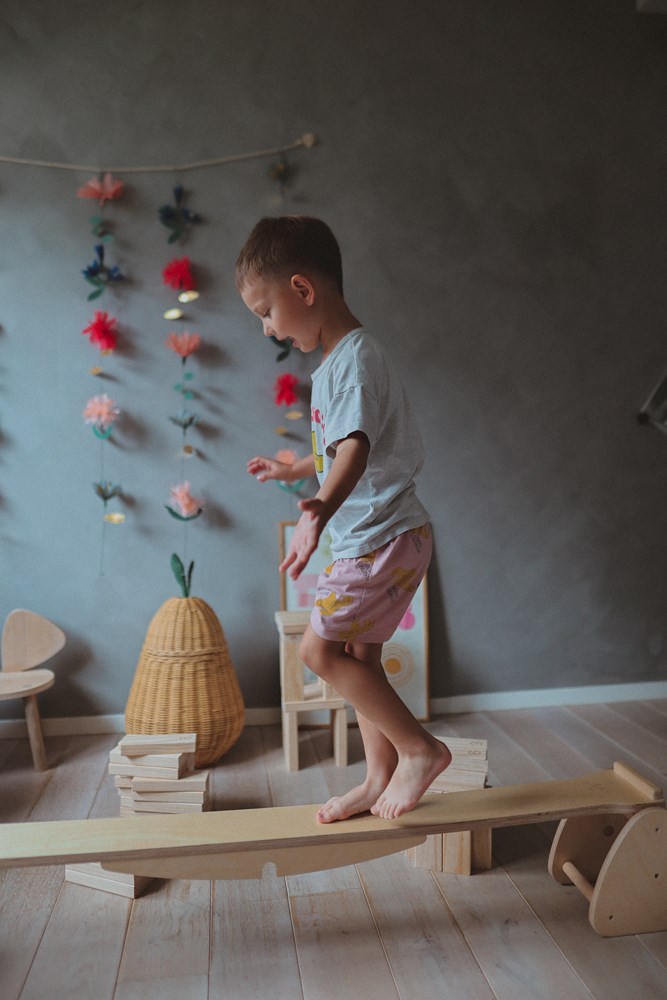 A child balances barefoot on the Kinderfeets Seesaw - Rocker in a cozy, flower-adorned room.