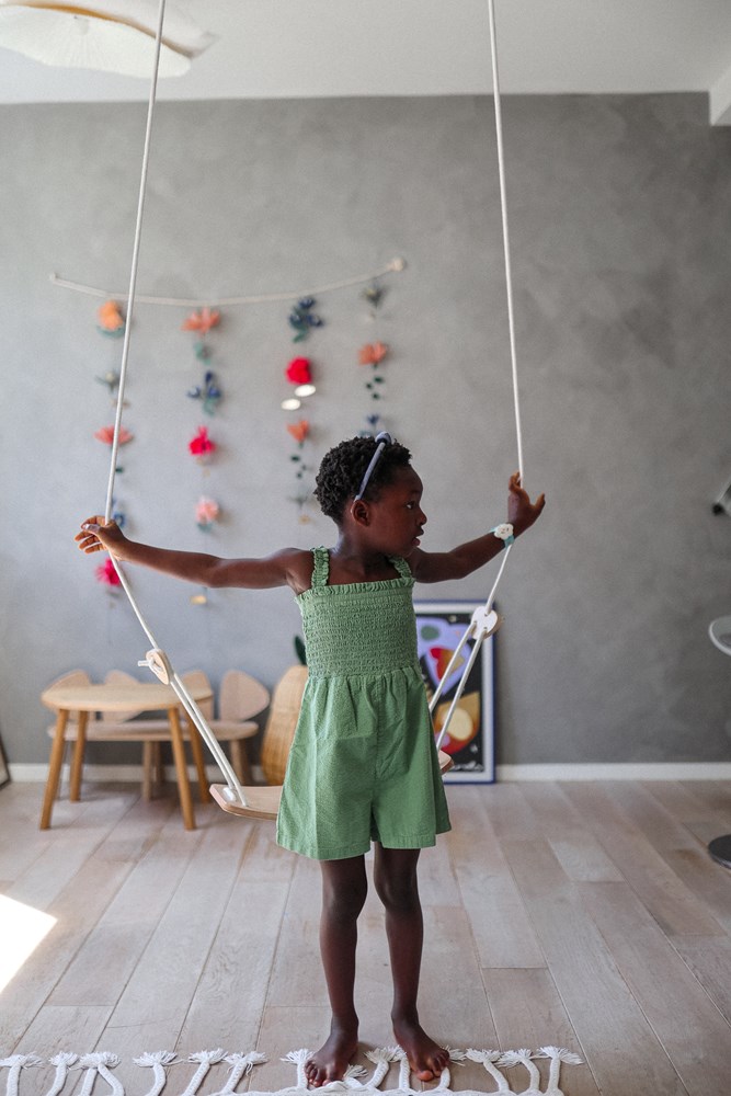 A small child stands on a "Kinderfeets" wooden swing in an indoor setting, with colorful wall decorations visible in the background.