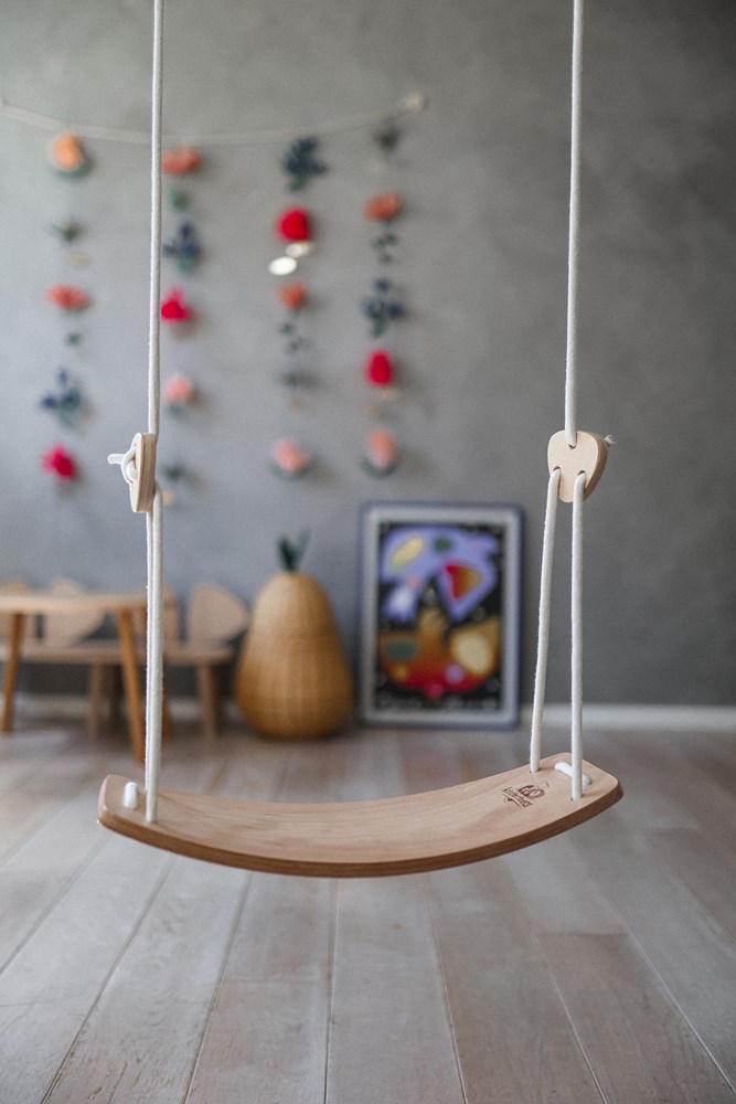 The "Kinderfeets" wooden swing hangs indoors, behind it on the floor are a wall decoration with a floral motif and a framed painting.