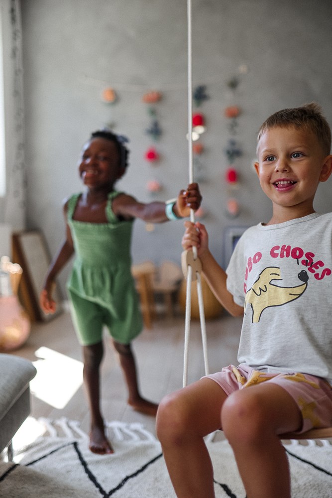 Two children are playing indoors; one is smiling on the "Kinderfeets Wooden Swing", the other is standing next to it – both are happy.