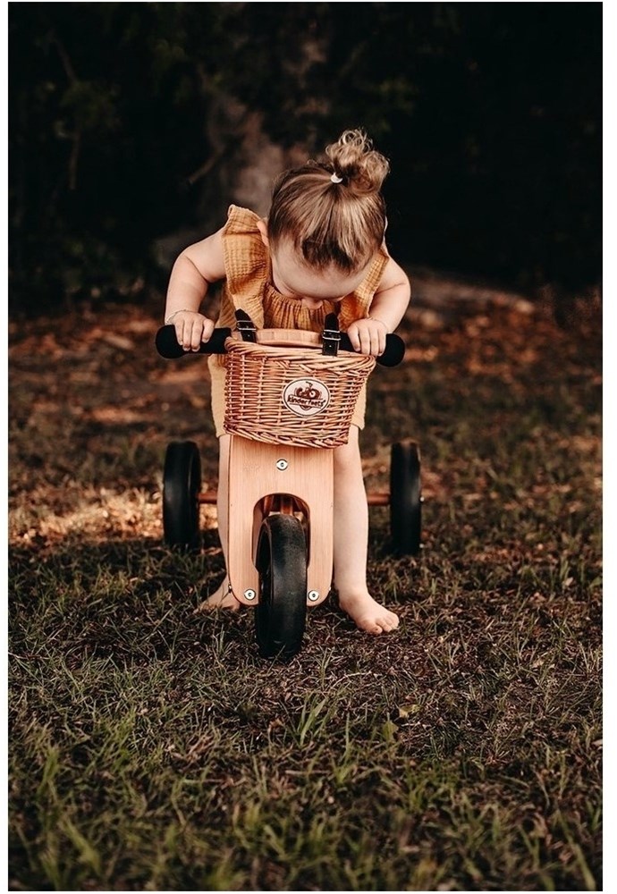 A small child is sitting outside on the lawn, and a "Kinderfeets" wicker bicycle basket is mounted on their wooden tricycle.
