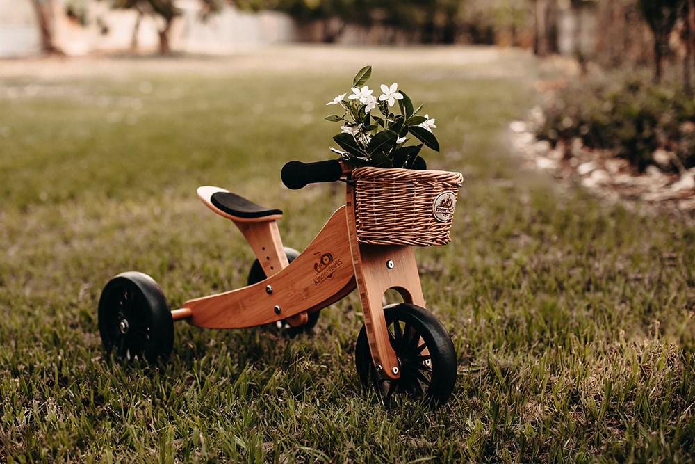 The Kinderfeets wicker bicycle basket with white flowers is on a wooden tricycle on the lawn in a park or garden.