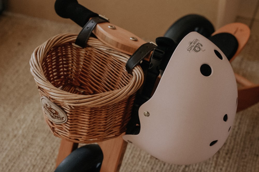 A Kinderfeets wicker bicycle basket and a white helmet are attached to the handlebars of a wooden balance bike indoors.
