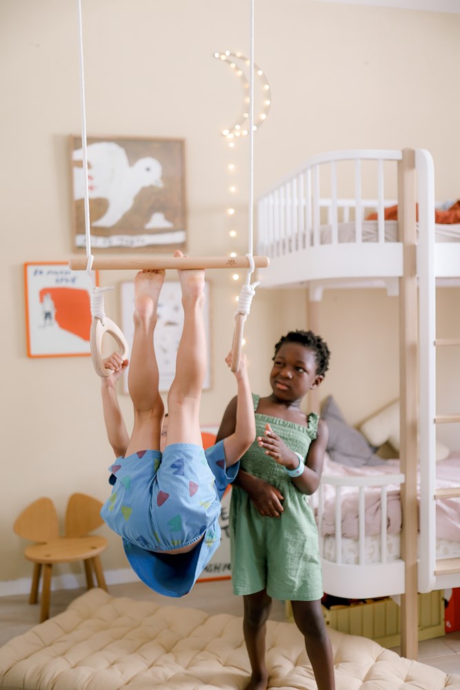 A child hangs from the “Kinderfeets Trapez mit Ringen” while another in a green dress stands beside it in a cozy bedroom.